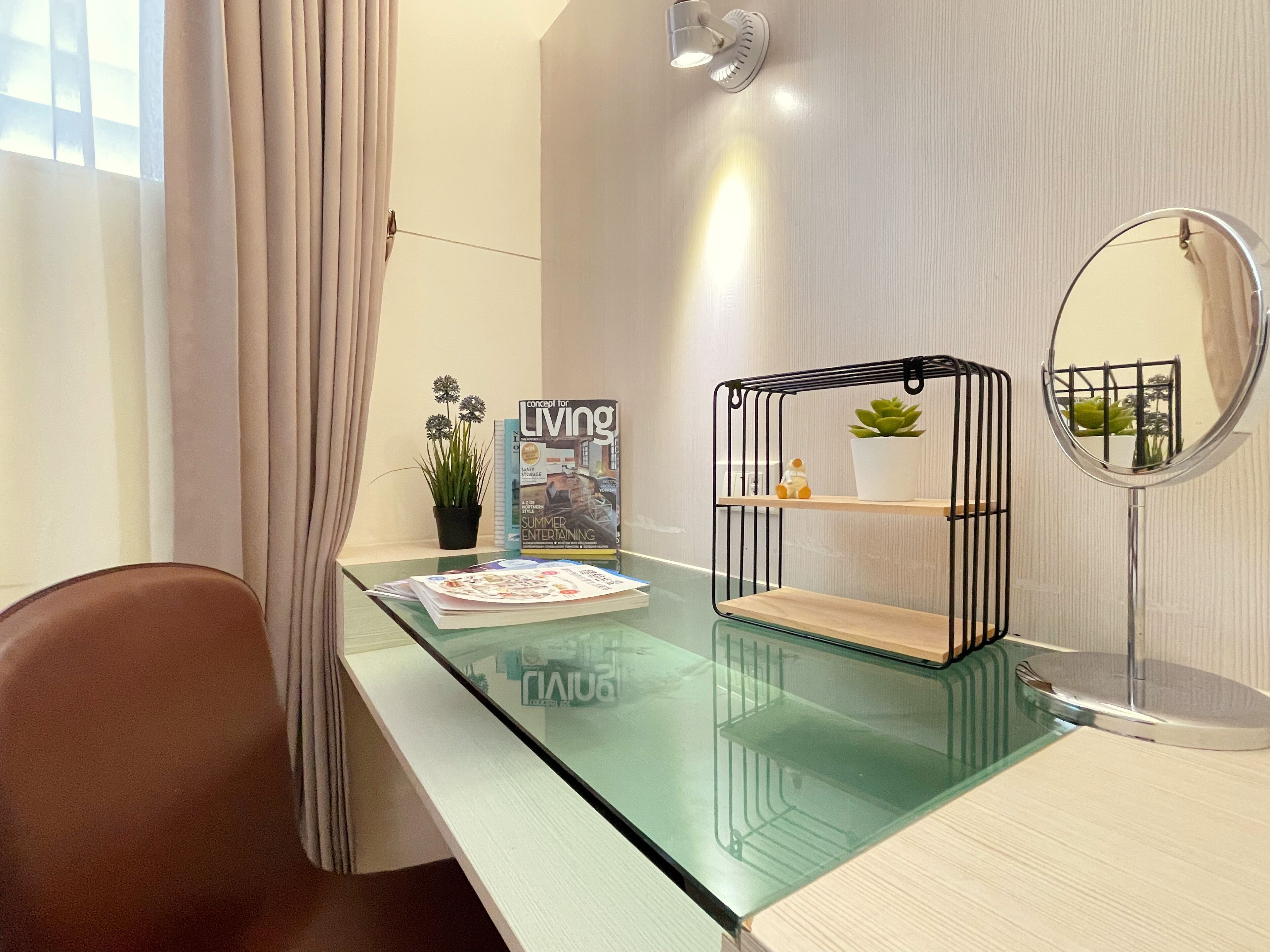 A modern room with a glass desk, a round mirror, a potted plant, a magazine, and a small shelf unit.