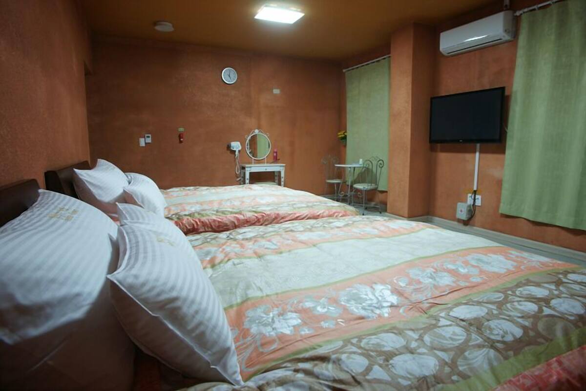 A bedroom with two beds, a bedside table with a mirror, a TV mounted on the wall, and an air conditioning unit.