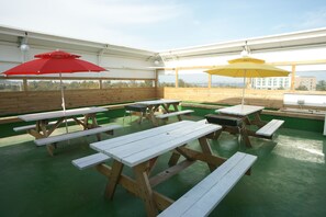 An outdoor patio area with picnic tables and umbrellas, wooden benches, and a view of the cityscape. - Feliz Town (Seogwipo)
