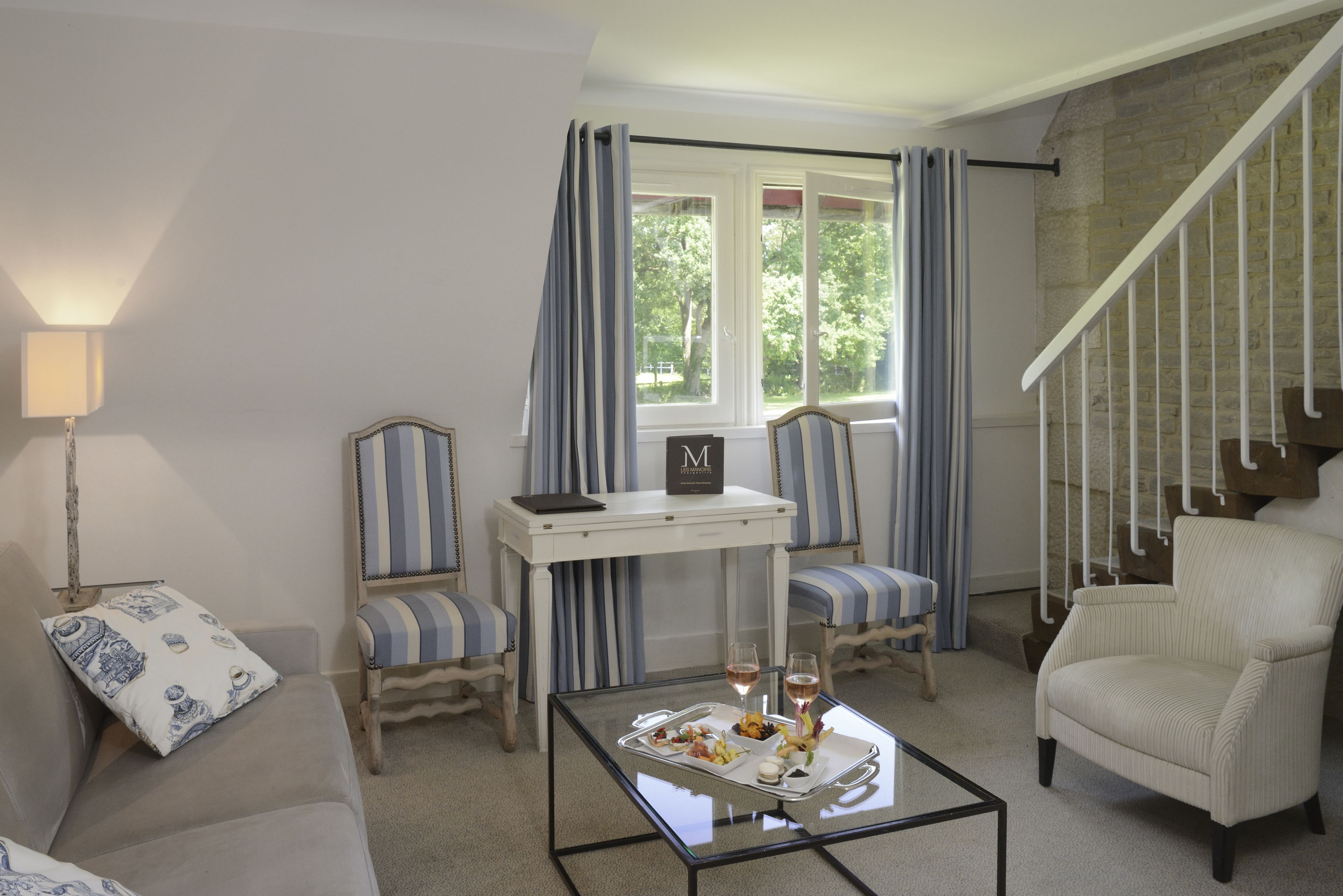 A living room with a sofa, two striped armchairs, a coffee table with a tray of food and drinks, a small table with a book, and a staircase with a stone wall.