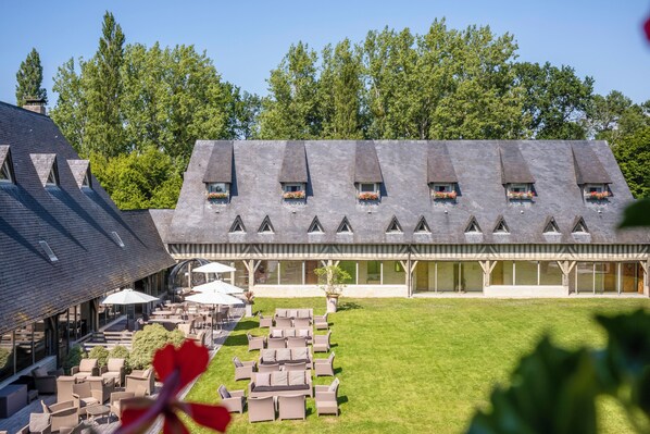 A building with multiple windows, a covered patio area with outdoor seating, and a grassy courtyard. - Les Manoirs de Tourgéville (Tourgeville)
