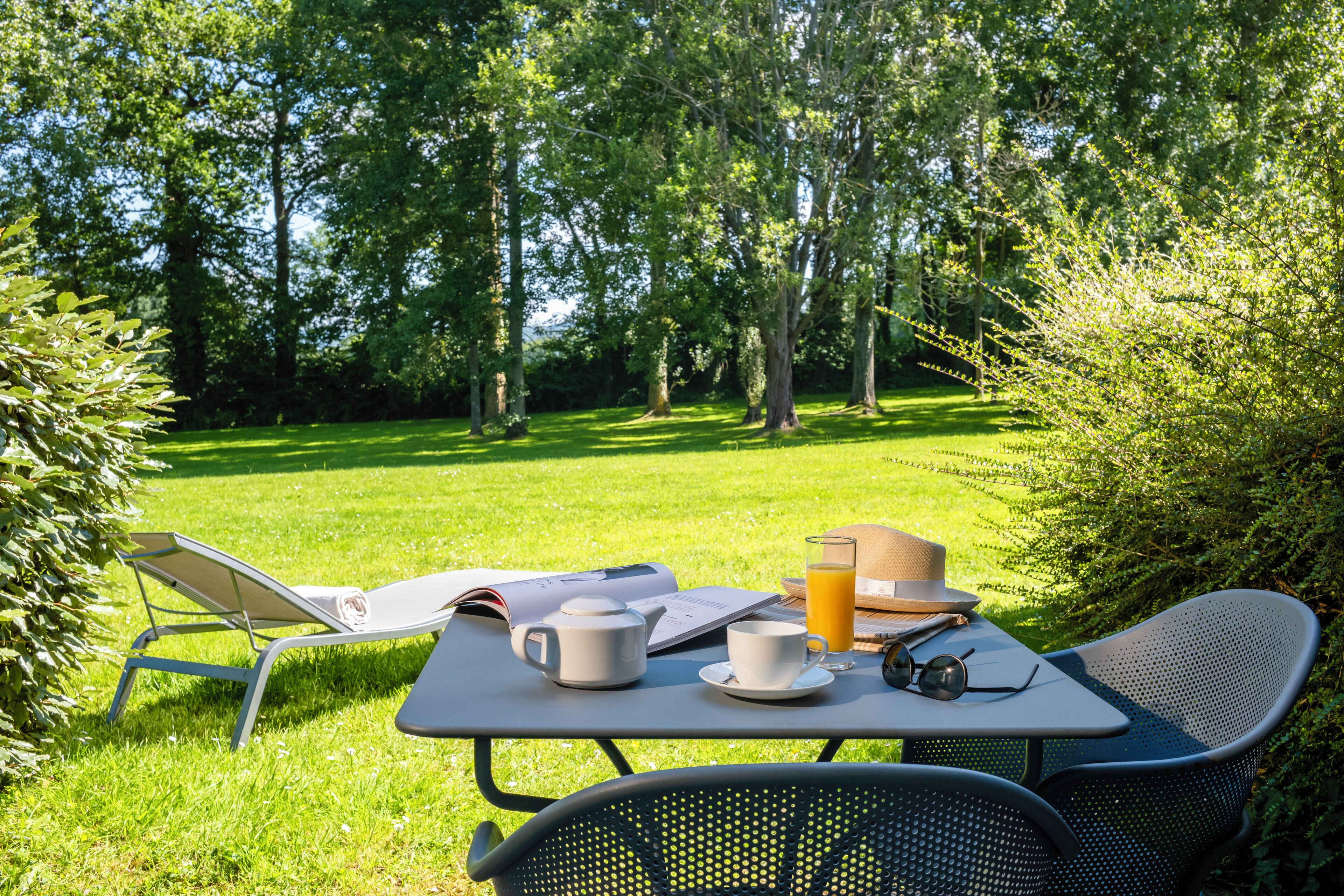 A table set outdoors with a book, a cup, a teapot, a glass of orange juice, and a hat.