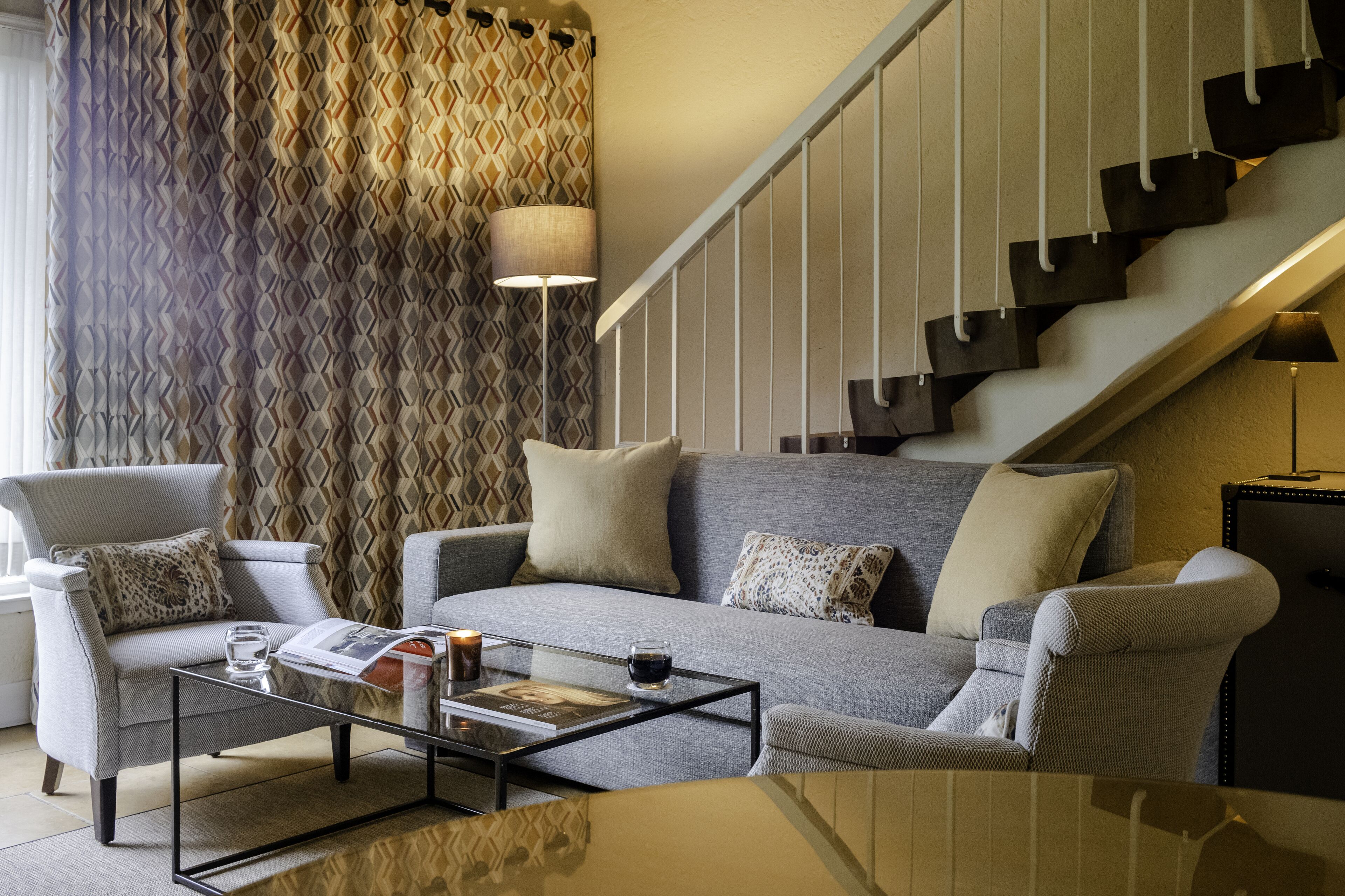 Una sala moderna con un sofá gris, dos sillones, una mesa de centro de vidrio y una escalera con un revestimiento de pared de estampado geométrico.