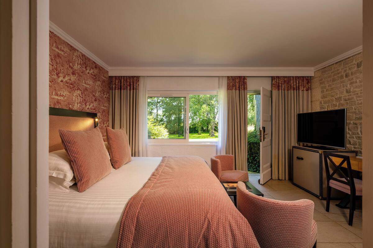 A hotel room with a large bed, a TV, a desk, and a view of greenery outside.