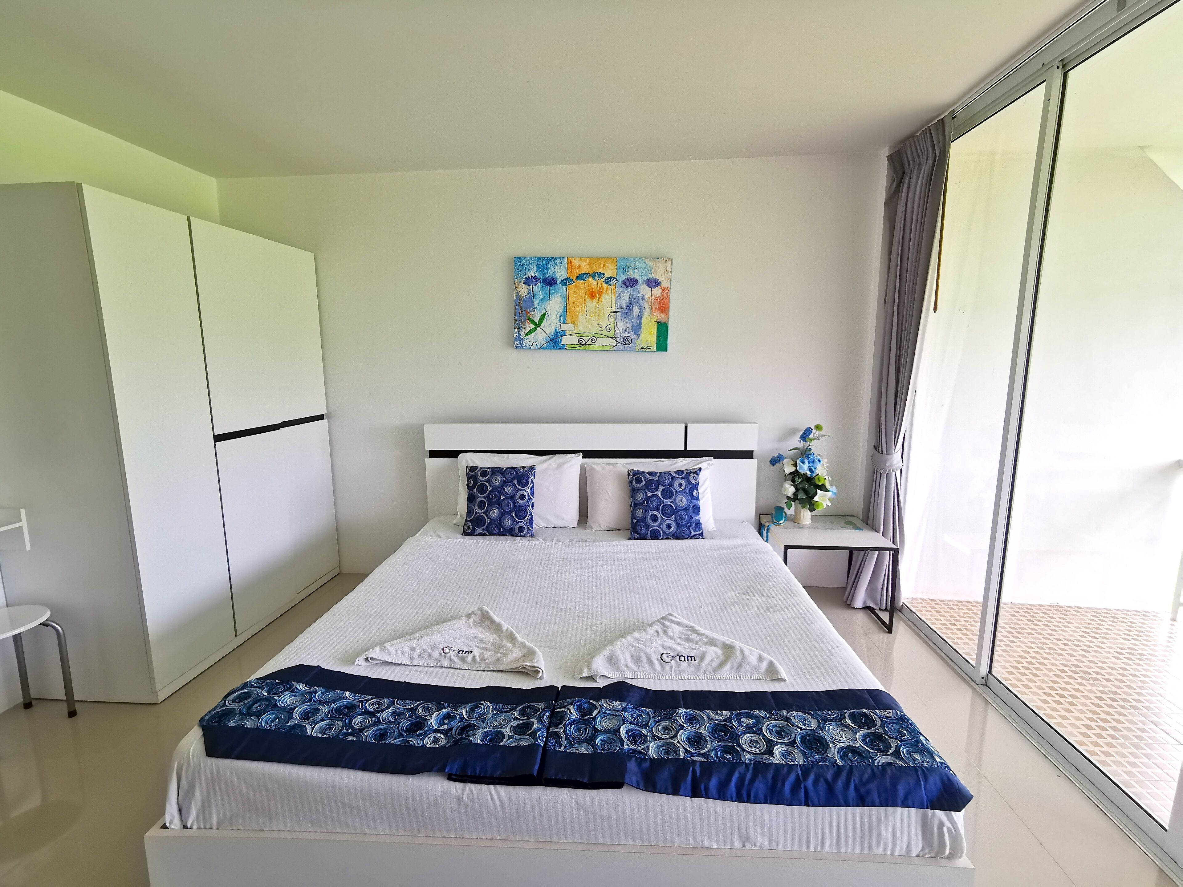 A modern hotel room with a large bed, a small bedside table with a vase of flowers, a painting on the wall, and a sliding glass door to the balcony.