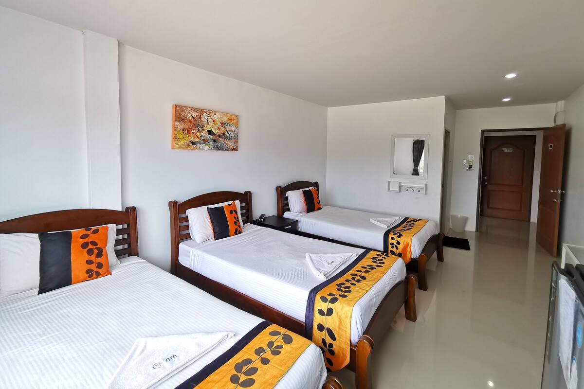 Habitación de hotel con tres camas individuales, cada una con colchón blanco y almohada decorativa anaranjada y negra. Sobre las camas hay un cuadro en la pared, un espejo y una puerta que da a otra habitación.