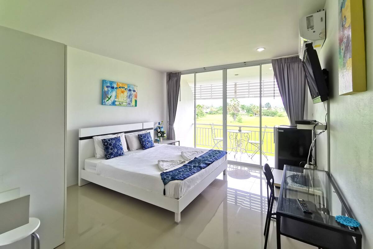 Habitación de hotel moderna con una cama grande, un escritorio con computadora, un televisor y un balcón con vista al jardín.