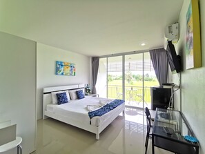 A modern hotel room with a large bed, a desk with a computer, a TV, and a balcony with a view of the garden.