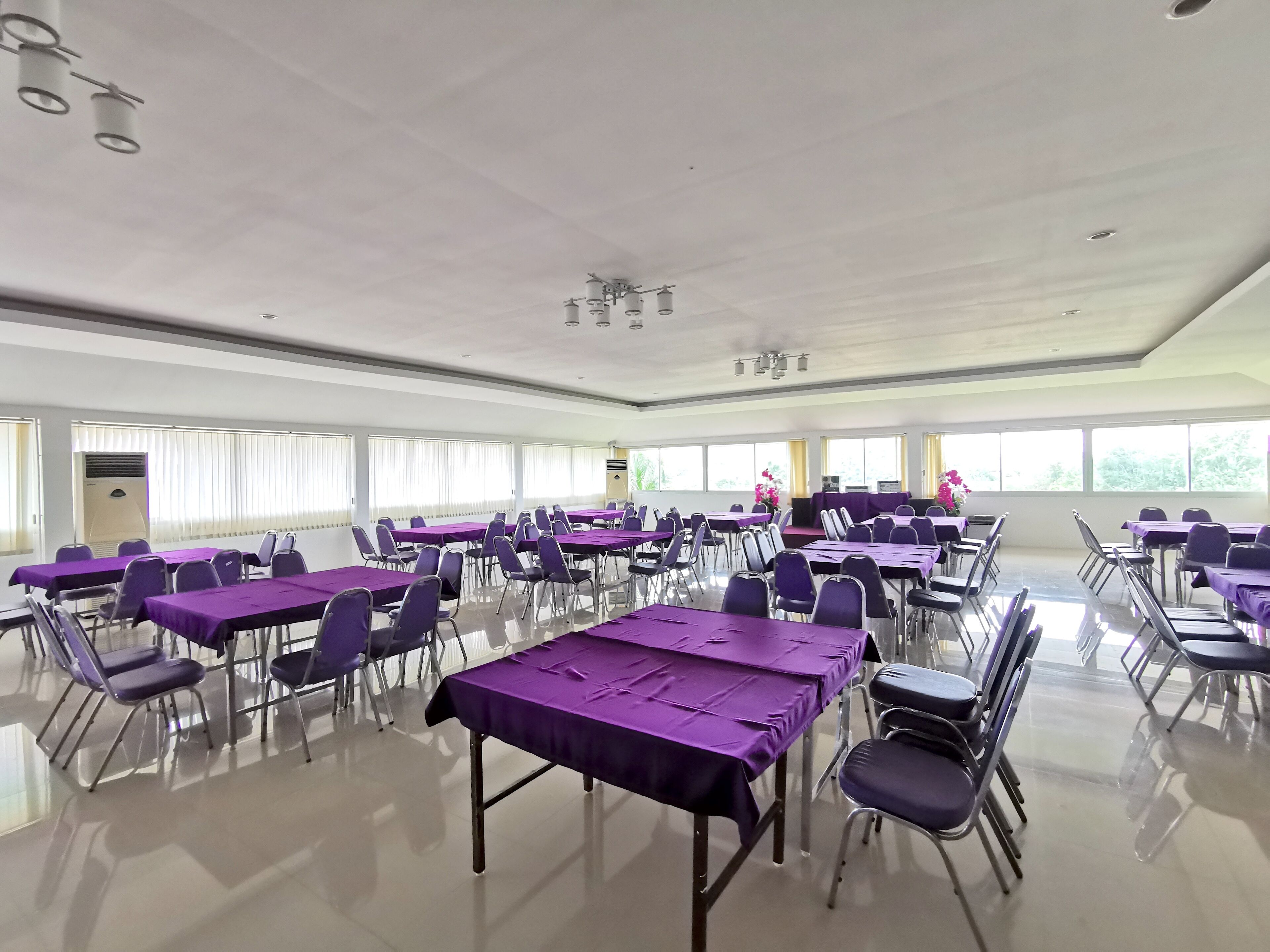 Un amplio salón de conferencias con filas de mesas y sillas, ventanas grandes y un escenario con atril.