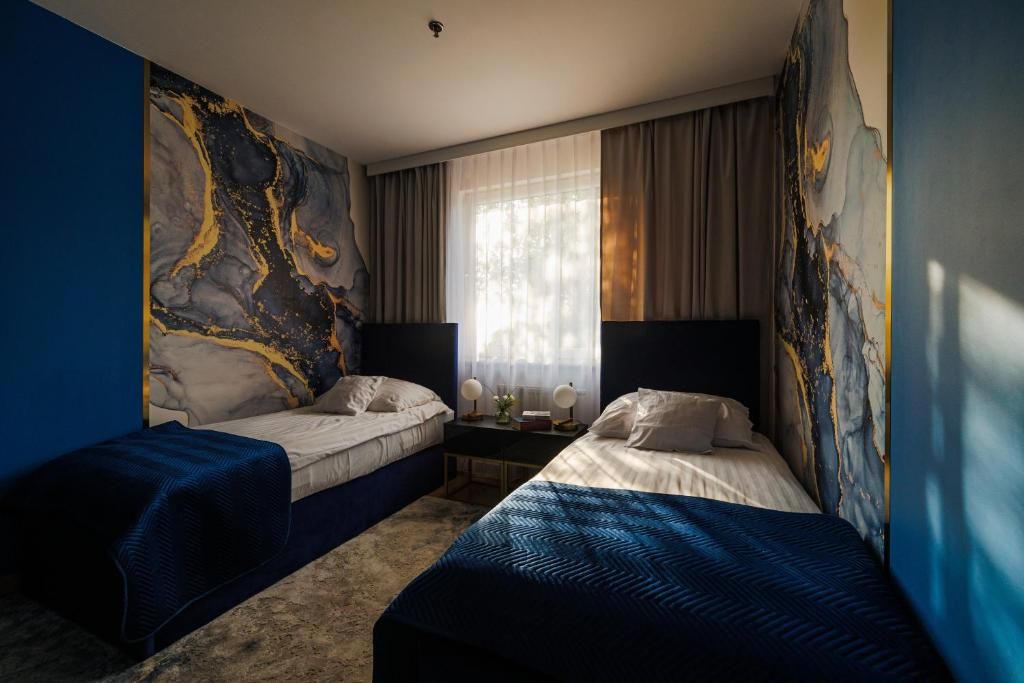 A hotel room with two beds, a large abstract painting on the wall, a window with curtains, and a nightstand with a lamp.