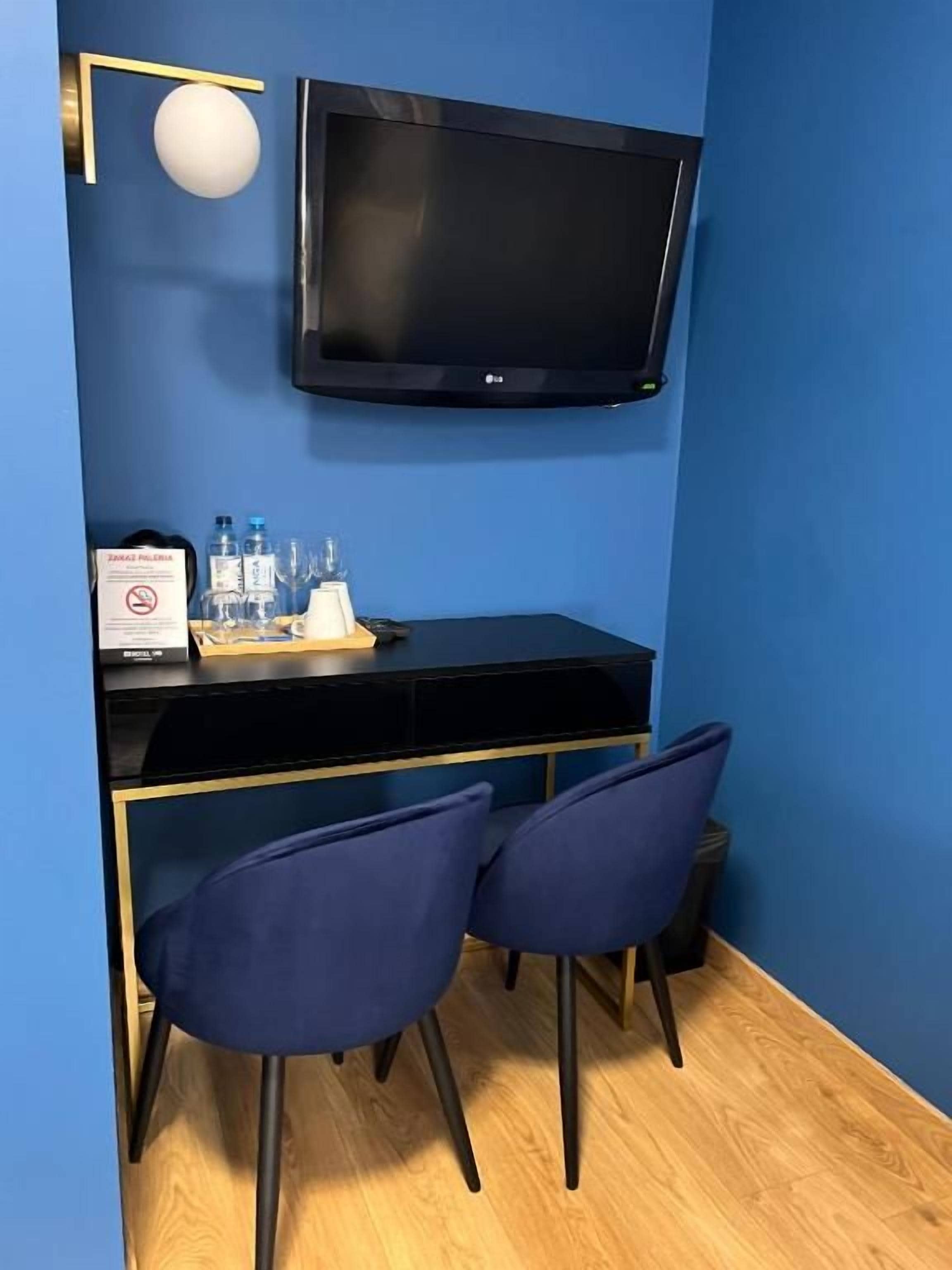 A room with a black desk, two blue chairs, a mounted TV, and a small table with water bottles and a sign.