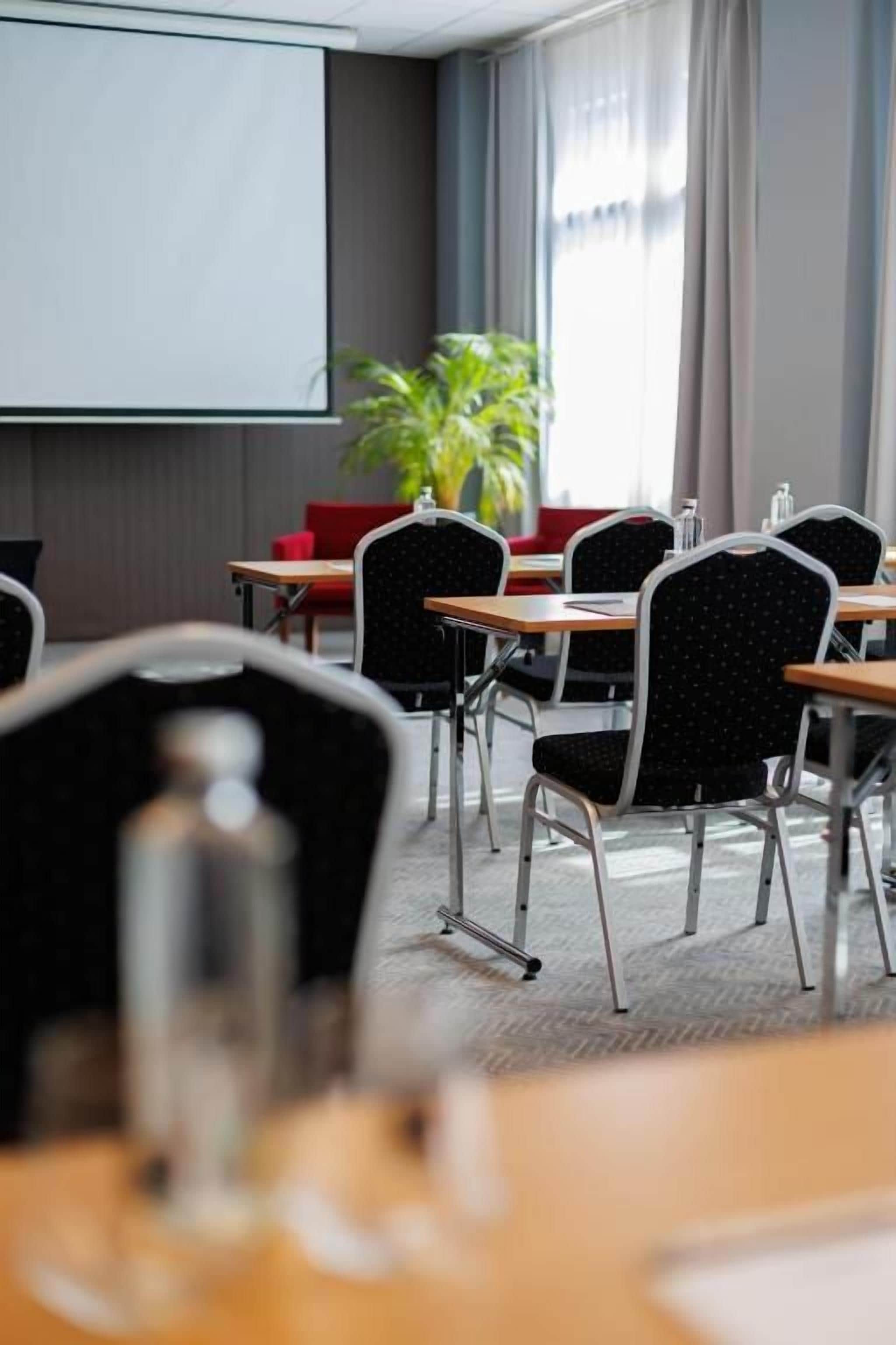 A conference room with a projector screen, tables, chairs, and a potted plant.