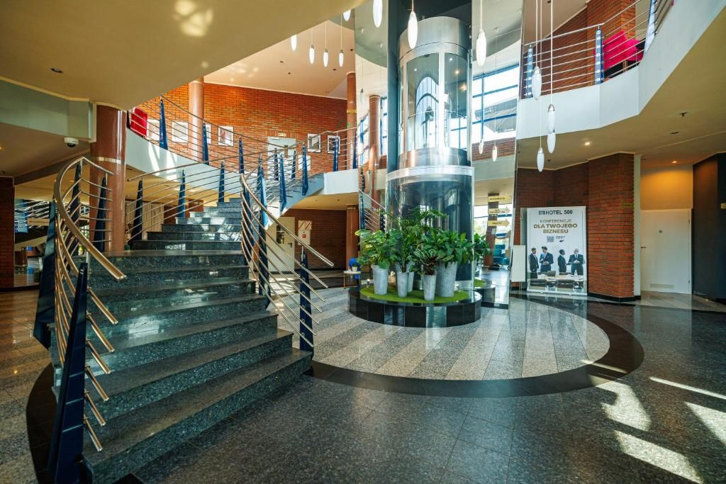 A hotel lobby with a central staircase, a circular column with plants, and a sign for HOTEL 500.