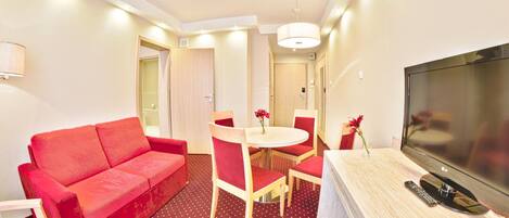A hotel room with a red sofa, a round dining table with red chairs, a TV, and a small table with a vase of flowers.
