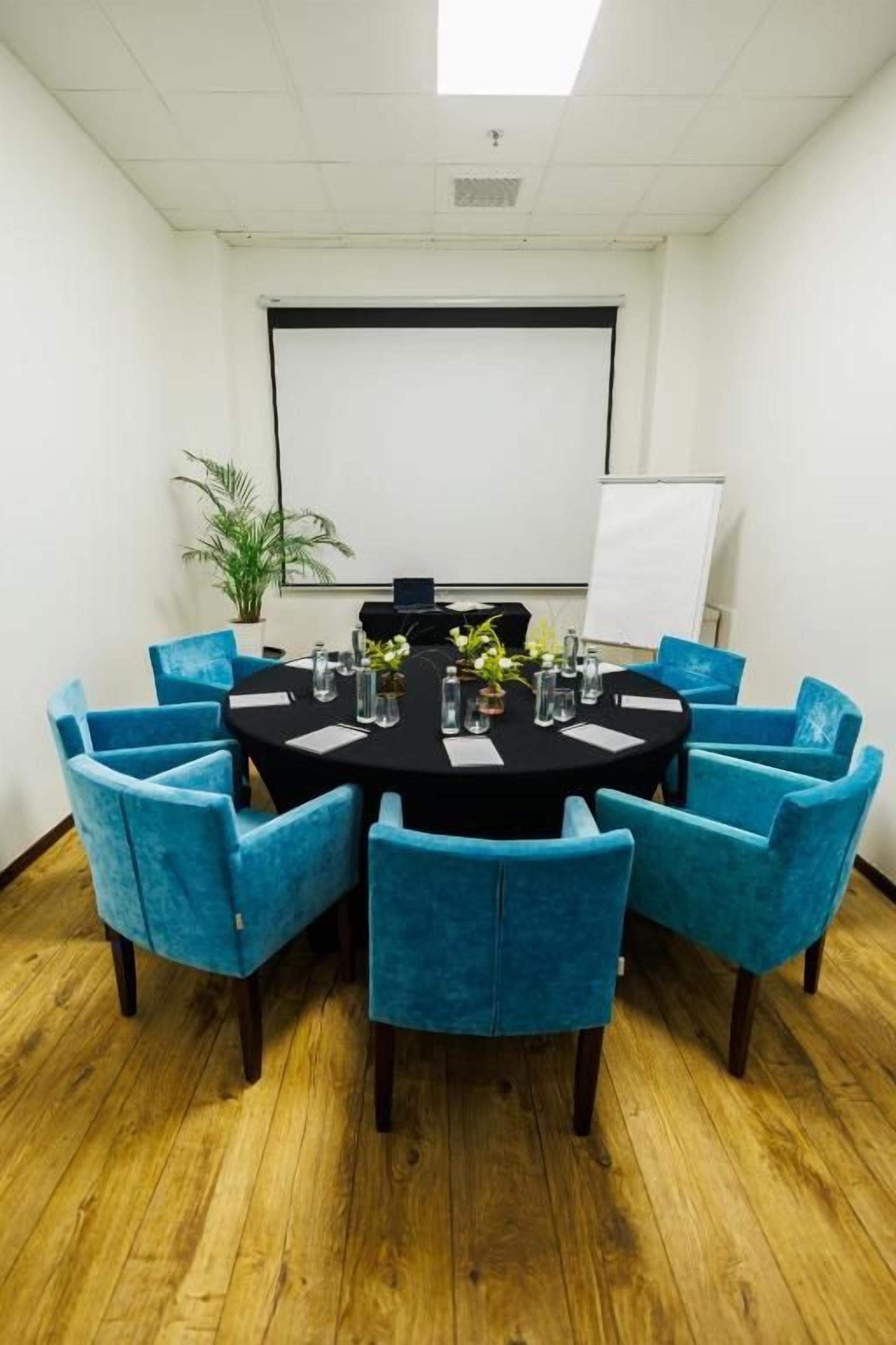 A conference room with a round table, blue chairs, a projector screen, and a whiteboard.