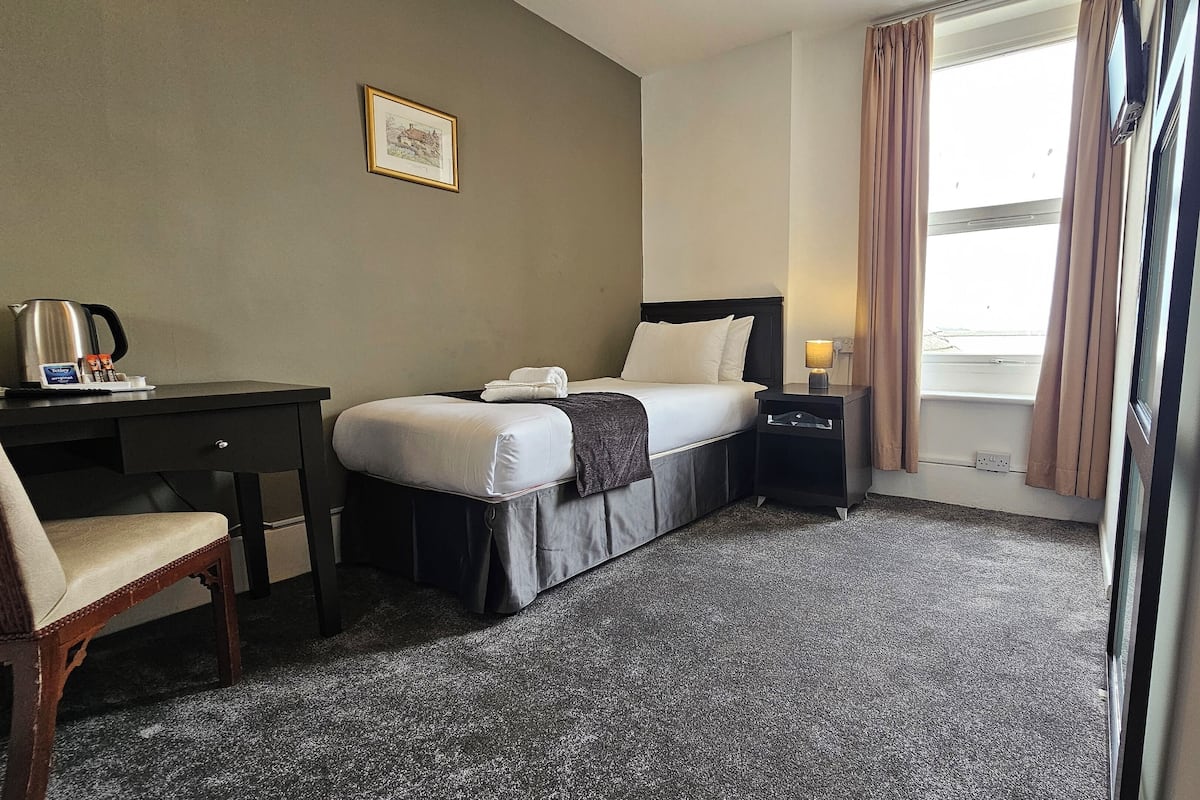 A hotel room with a bed, a desk, a chair, a nightstand, a lamp, a picture on the wall, and a window with curtains.