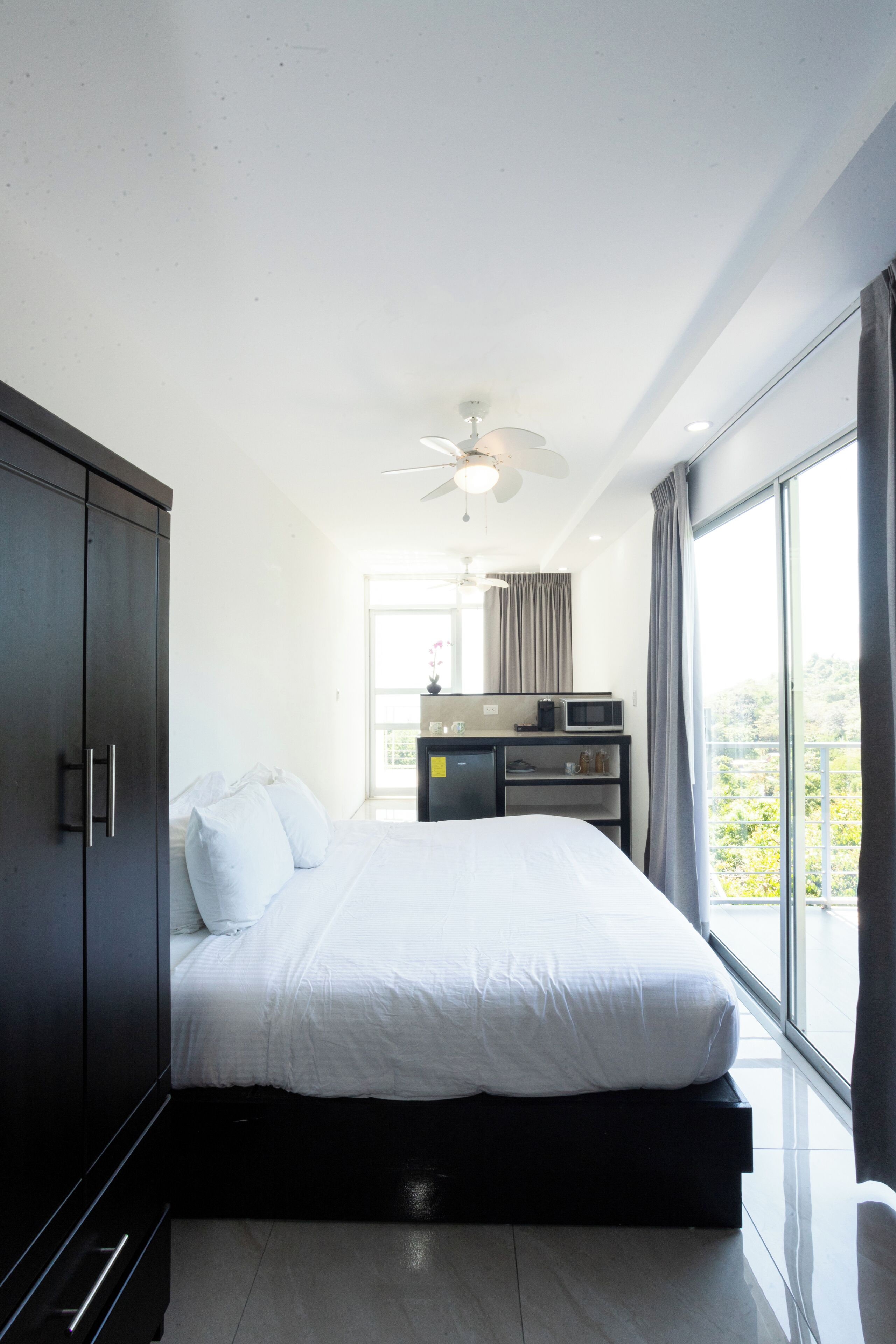 A modern hotel room with a large bed, a wardrobe, a desk with a computer, and a view of greenery through the window.