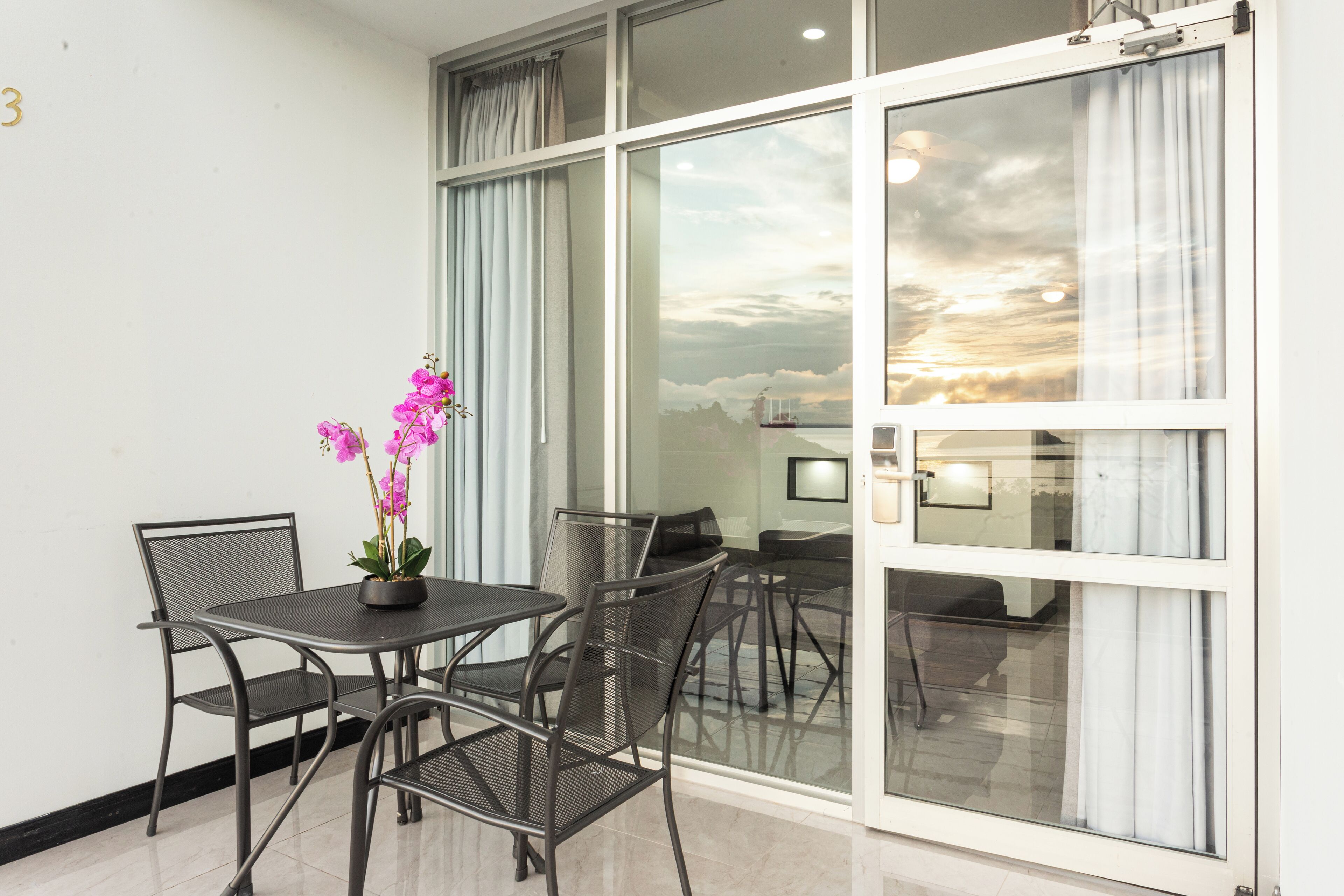 Une chambre moderne dotée d’une porte vitrée donnant sur un balcon, d’une table à manger avec des chaises et d’une plante orchidée en pot.