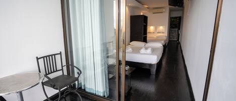 Habitación de hotel con puerta de vidrio que da a un balcón, cama con ropa blanca, una mesita con una silla y televisor montado en la pared.