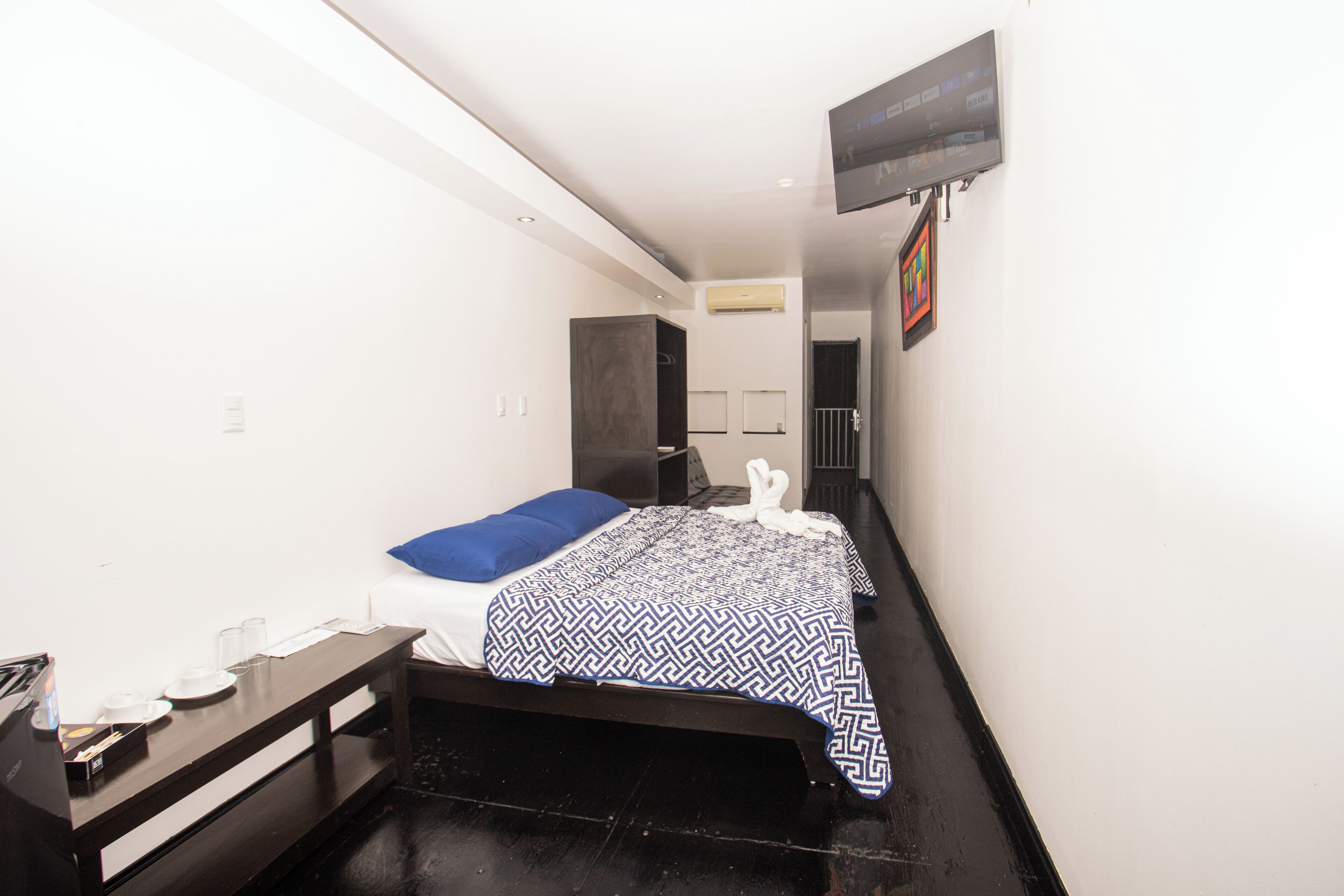A small, modern hotel room with a single bed, a bedside table with a lamp, a flat-screen TV mounted on the wall, and a small window.