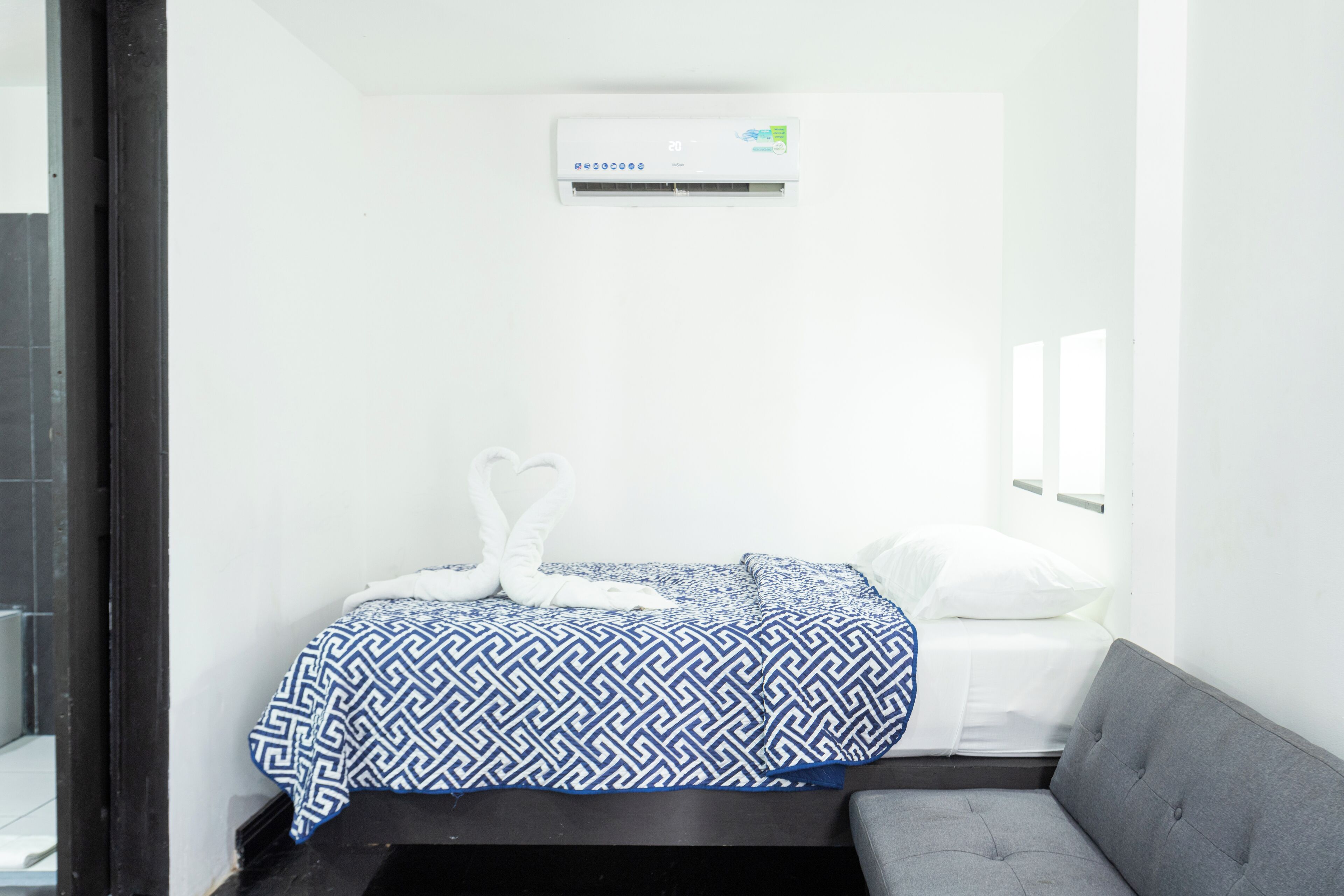 A small, clean room with a bed, a gray sofa, and a wall-mounted air conditioner.