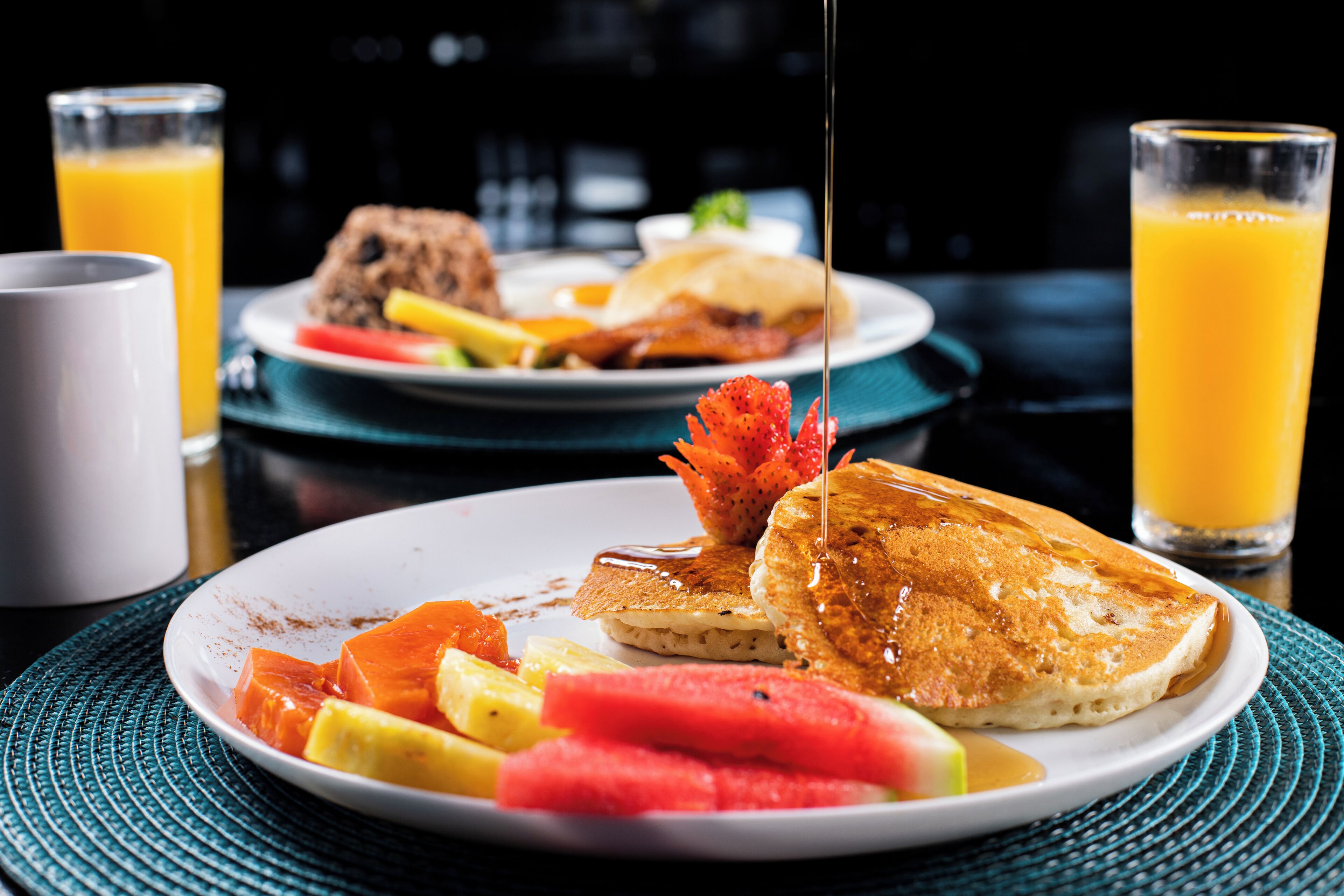 Une assiette de petit-déjeuner avec des crêpes, des fruits et du sirop, ainsi que deux verres de jus d’orange, posés sur une table.