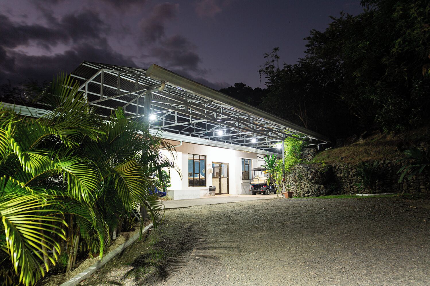 A modern building with a metal roof structure, surrounded by lush greenery and a gravel driveway.
