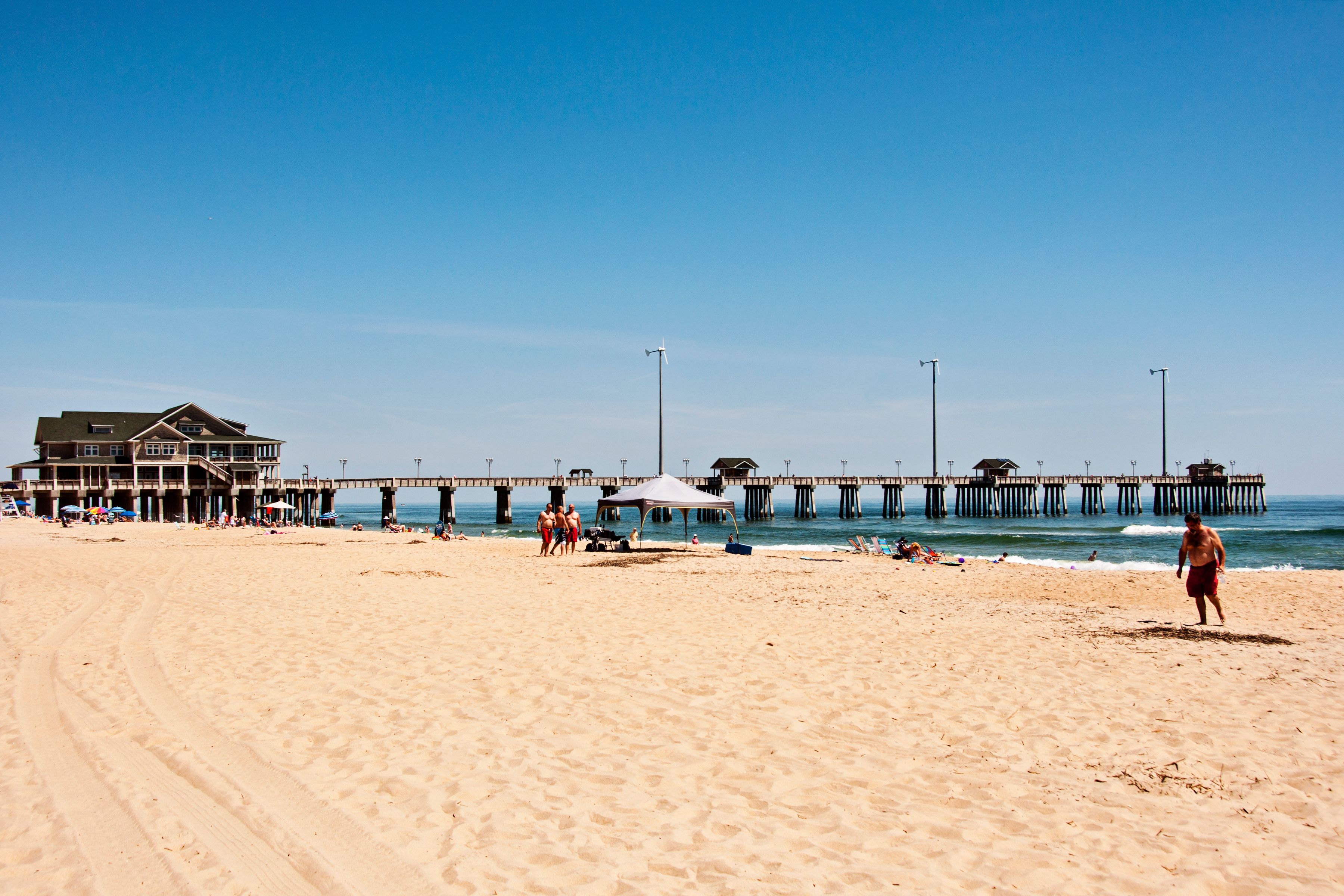 A beach with a pier extending into the water, a building with a balcony overlooking the beach, and people walking along the shore.
