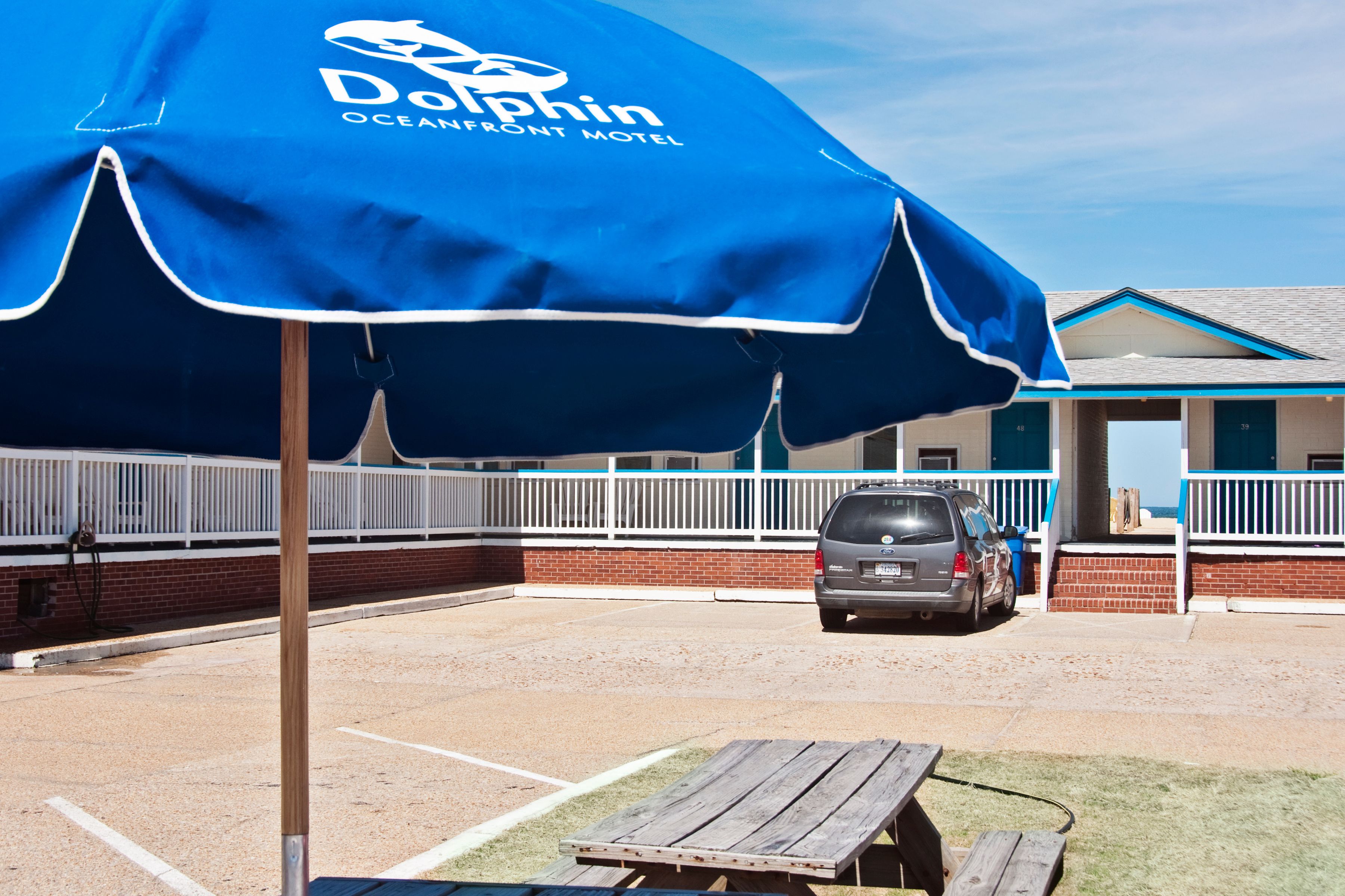 A blue Dolphin Oceanfront Motel umbrella, a parking area, and a building with a brick facade.