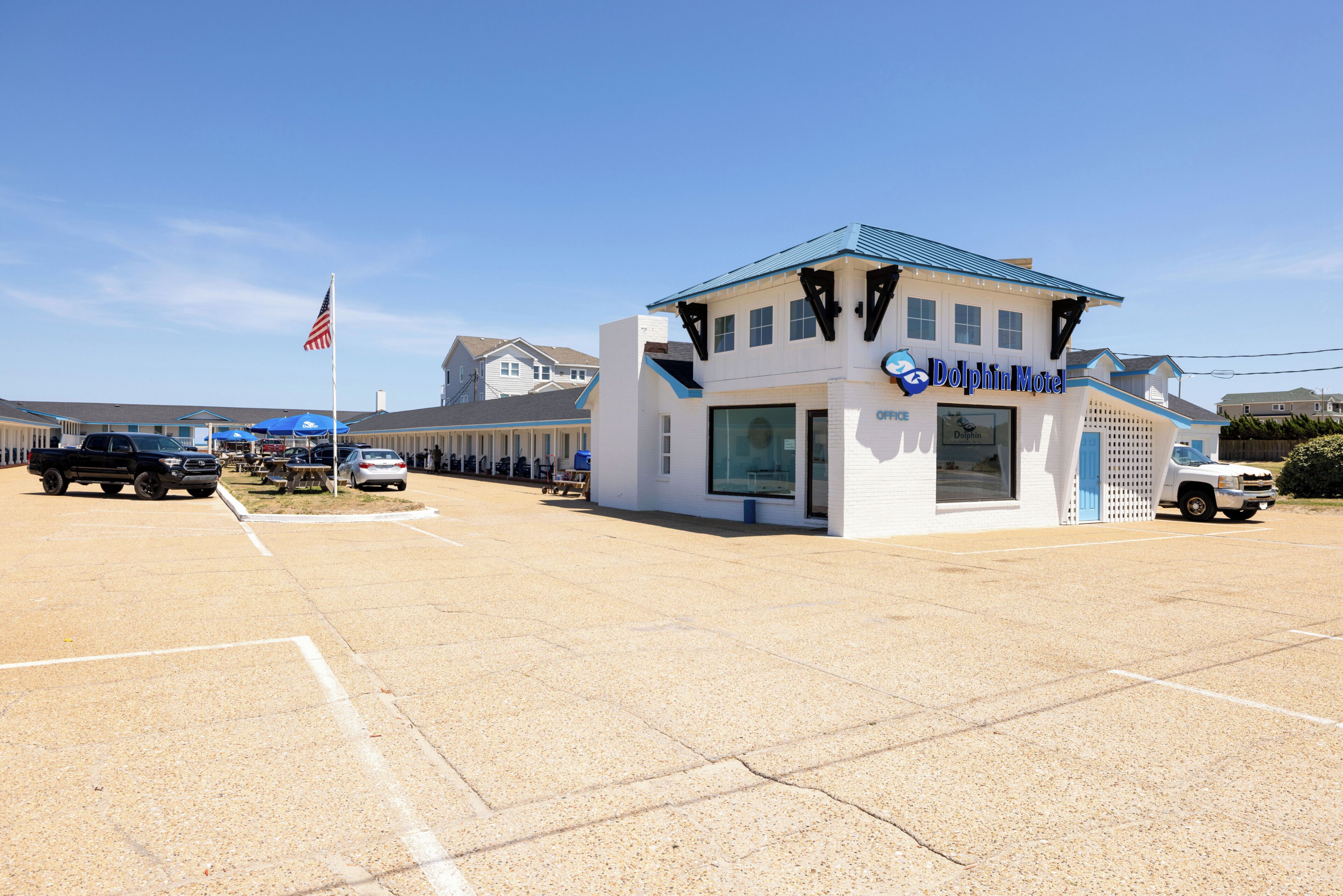 A Dolphin Motel with a white exterior and blue accents, a parking lot with several vehicles, and a clear sky.
