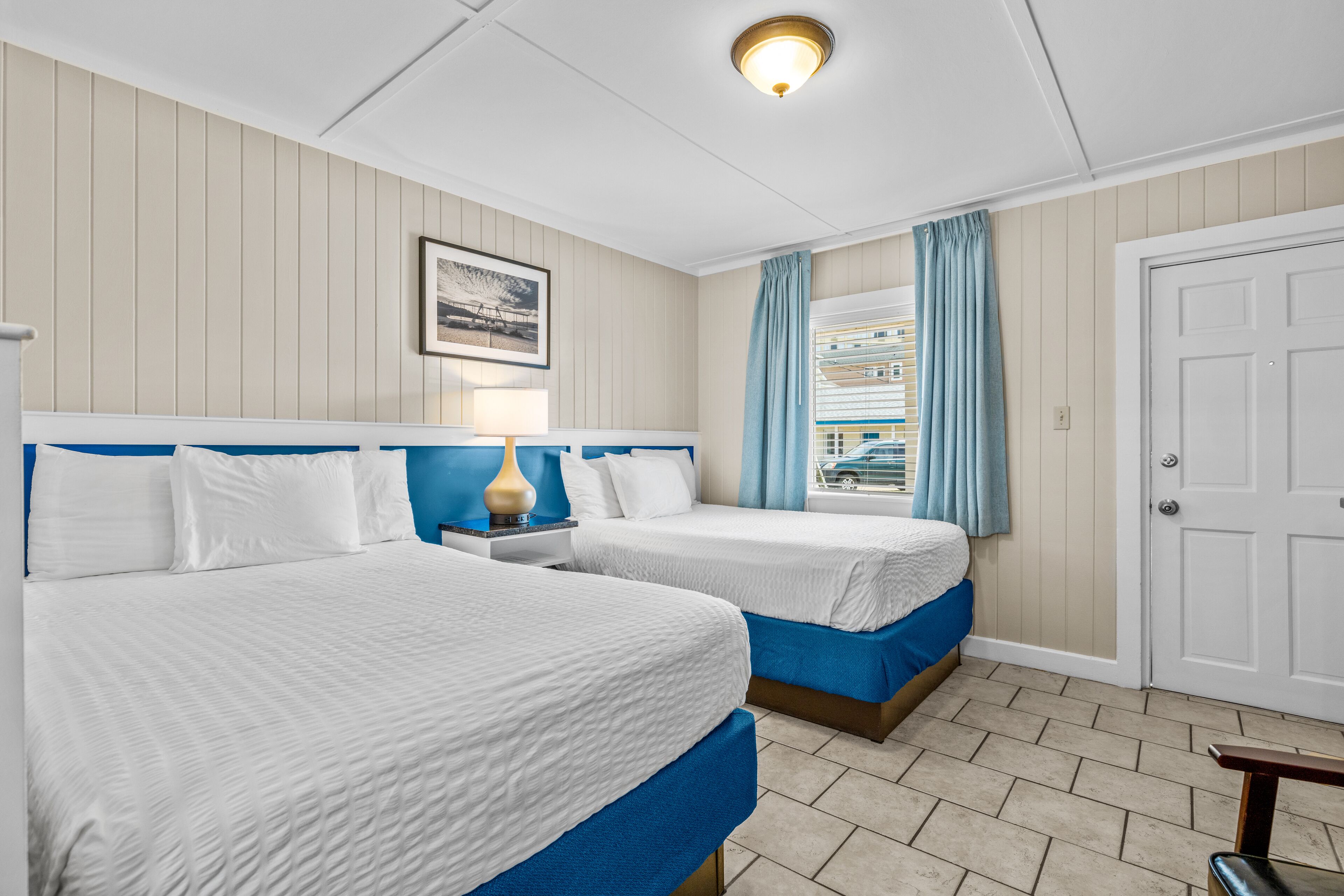 A hotel room with two beds, a nightstand, a lamp, a framed picture on the wall, and a window with curtains.
