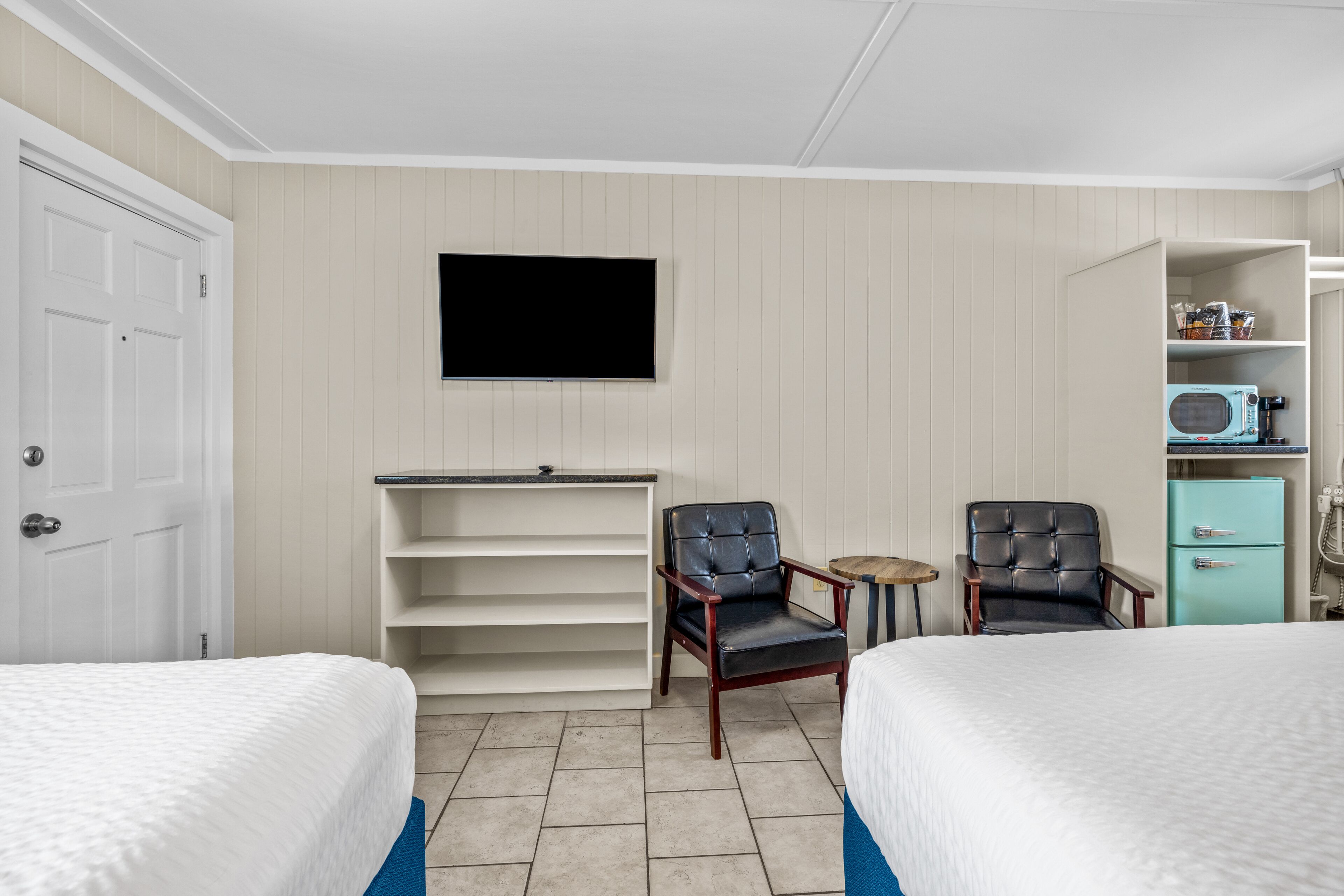 A hotel room with two beds, a television mounted on the wall, a small table, a chair, and a kitchenette with a microwave.