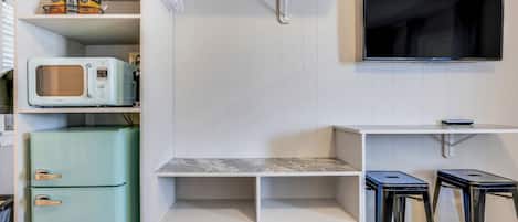 A compact kitchen with a built-in microwave, a small refrigerator, and a flat-screen TV mounted on the wall.
