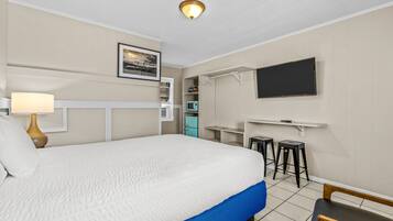 A hotel room with a large bed, a desk with a chair, a TV mounted on the wall, and a small kitchenette area.