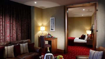 Een hotelkamer met een leren bank, een houten bureau met een lamp, een bed met een rode deken en een schaal fruit op de salontafel.