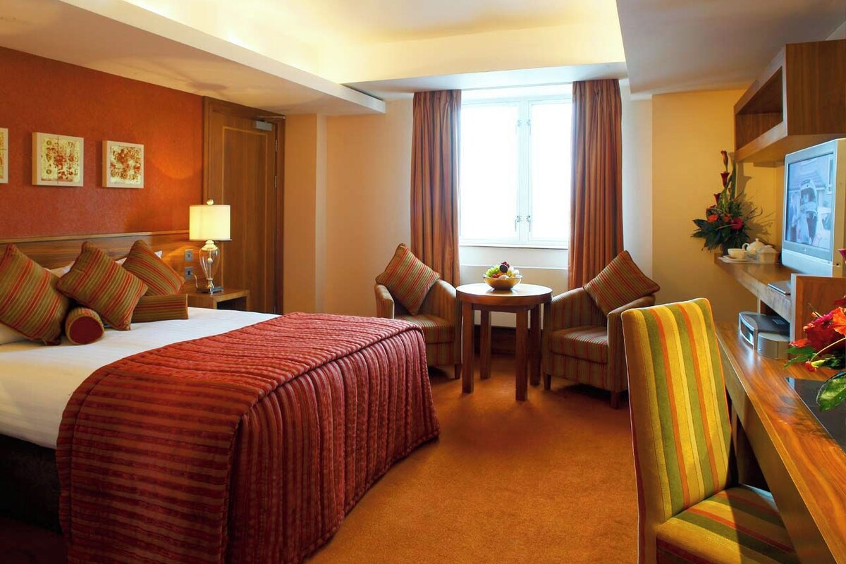 Habitación de hotel con cama, escritorio con televisor, silla, mesita con frutero y ventana con cortinas.