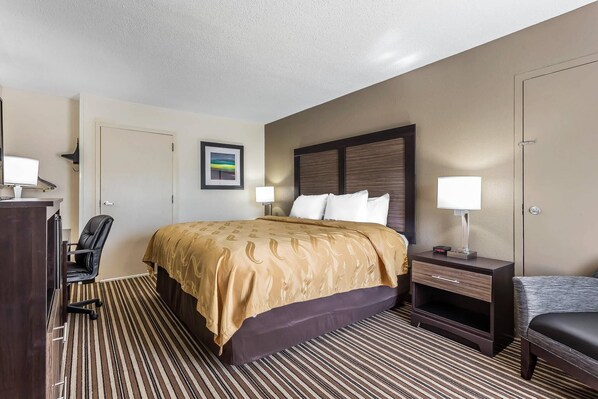 A hotel room with a bed, a desk, a chair, a nightstand with a lamp, and a picture on the wall. - Quality Inn Pinehurst Area (Aberdeen)