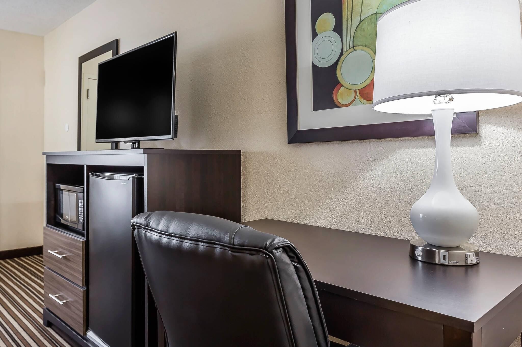 Habitación de hotel con escritorio, silla, televisor, microondas, refrigerador y lámpara.