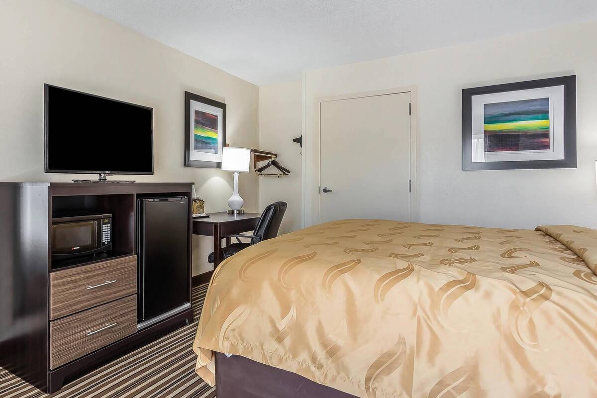 A hotel room with a bed, a TV, a desk with a lamp, a microwave, and a refrigerator.