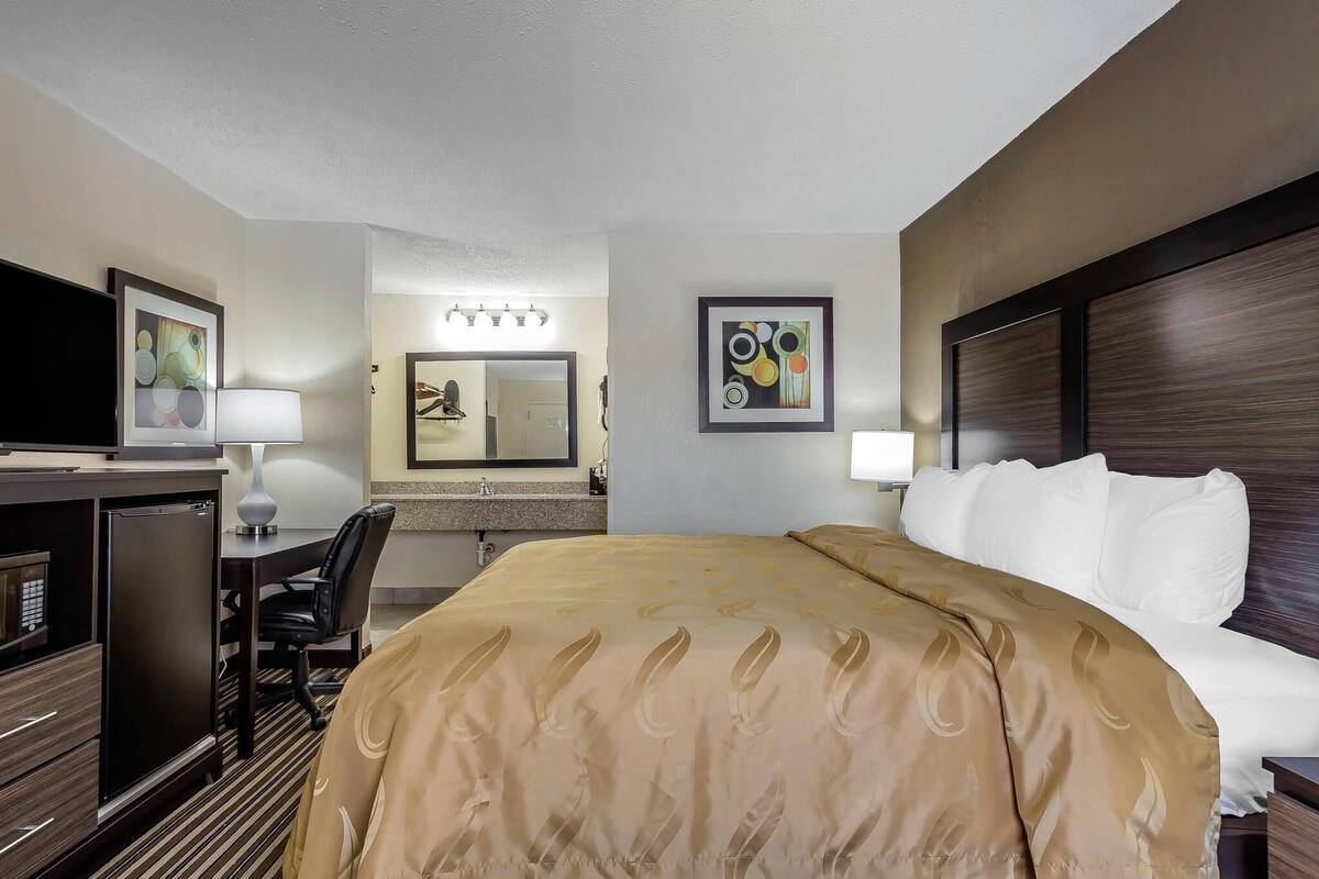 A hotel room with a bed, a desk with a computer, a chair, a mirror, and artwork on the walls.