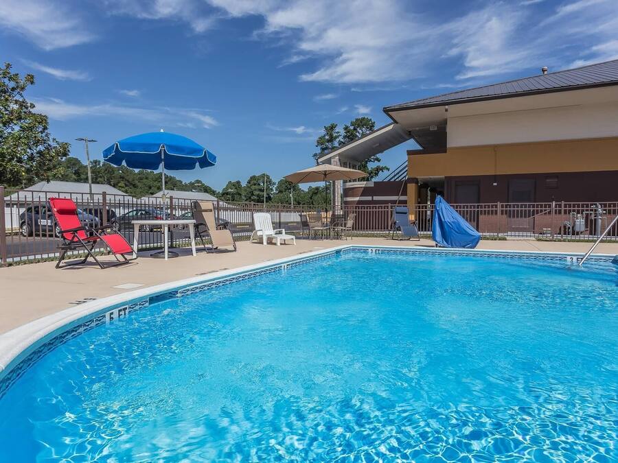 A swimming pool with lounge chairs and umbrellas, surrounded by a fenced area and a building with a flat roof.