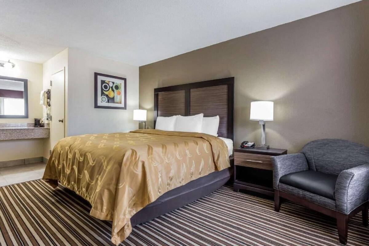 A hotel room with a bed, a chair, a nightstand, a lamp, a painting, and a bathroom with a mirror and sink.