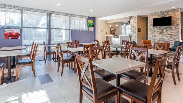 A modern dining area with wooden tables and chairs, large windows with curtains, and a stone accent wall with a flat-screen TV. - Quality Inn Pinehurst Area (Aberdeen)