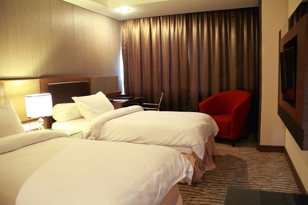 Une chambre d’hôtel avec deux lits, une chaise rouge, un bureau avec une lampe et une grande fenêtre avec des rideaux.
