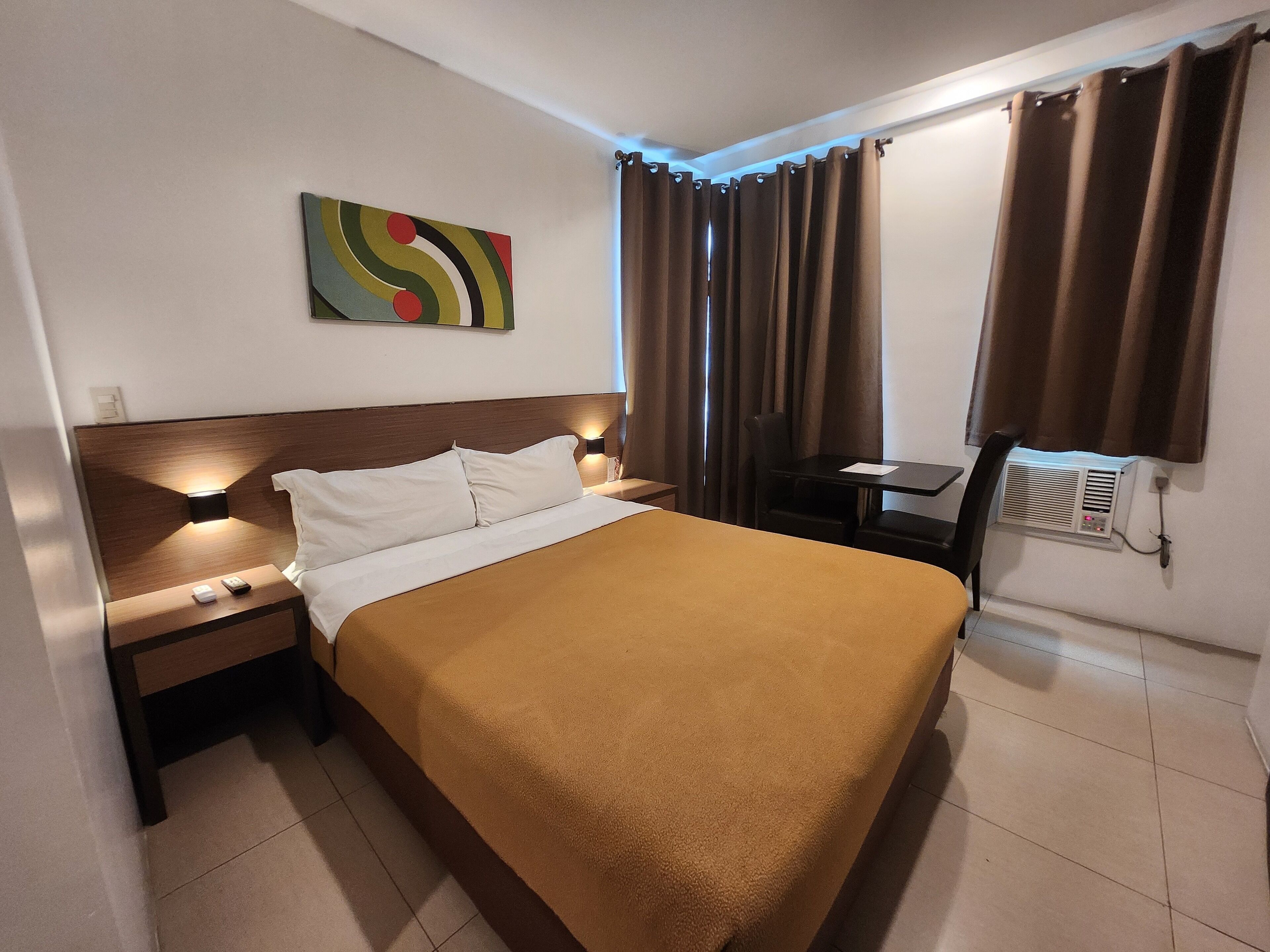 A hotel room with a bed, a desk, a chair, a wall-mounted air conditioner, and a colorful abstract painting.