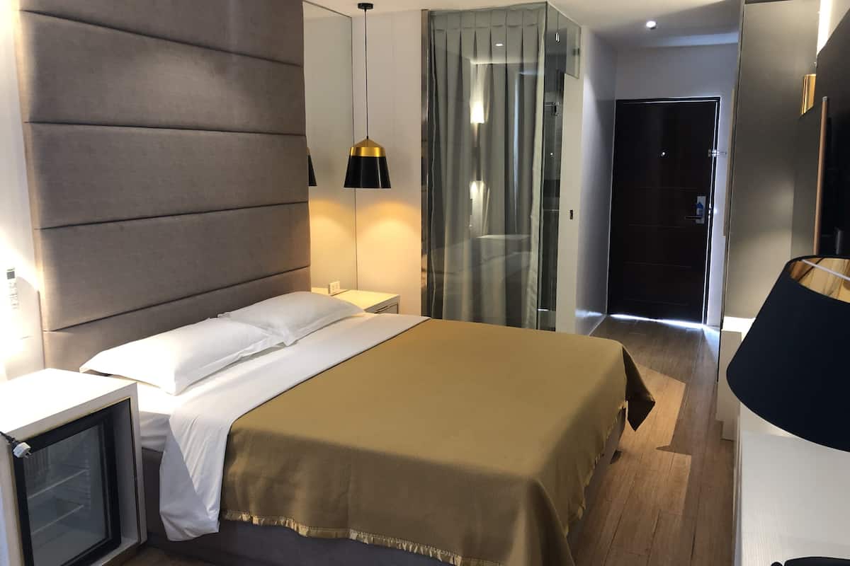 A modern hotel room with a large bed, bedside tables, a nightstand, a lamp, and a glass partition.