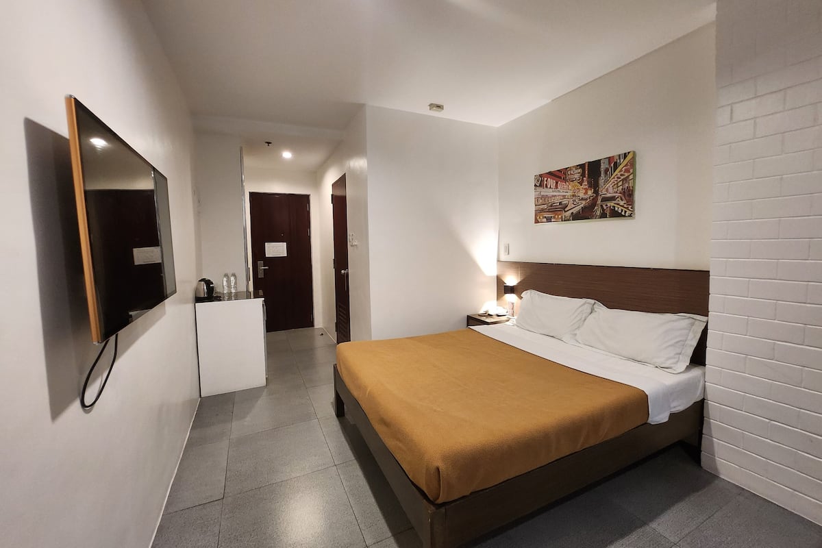 A hotel room with a bed, a flat-screen TV, a desk, and a door leading to a hallway.