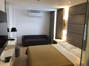 A hotel room with a bed, a sofa, a desk with a lamp, a mirror, and an air conditioning unit. - Apollonia Royale Hotel (Angeles City)