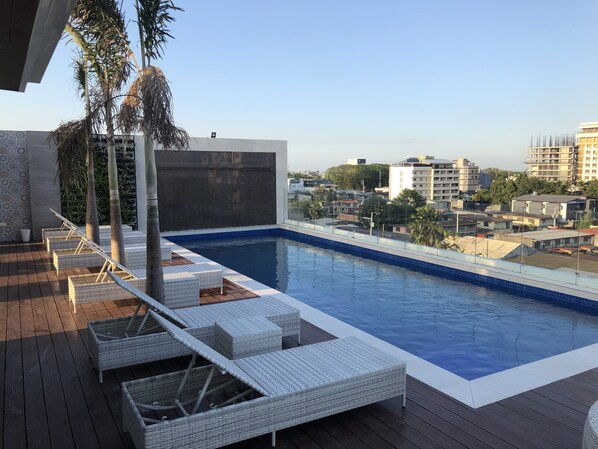 A rooftop pool with white wicker loungers, a clear view of the cityscape, and a modern building in the background. - Apollonia Royale Hotel (Angeles City)
