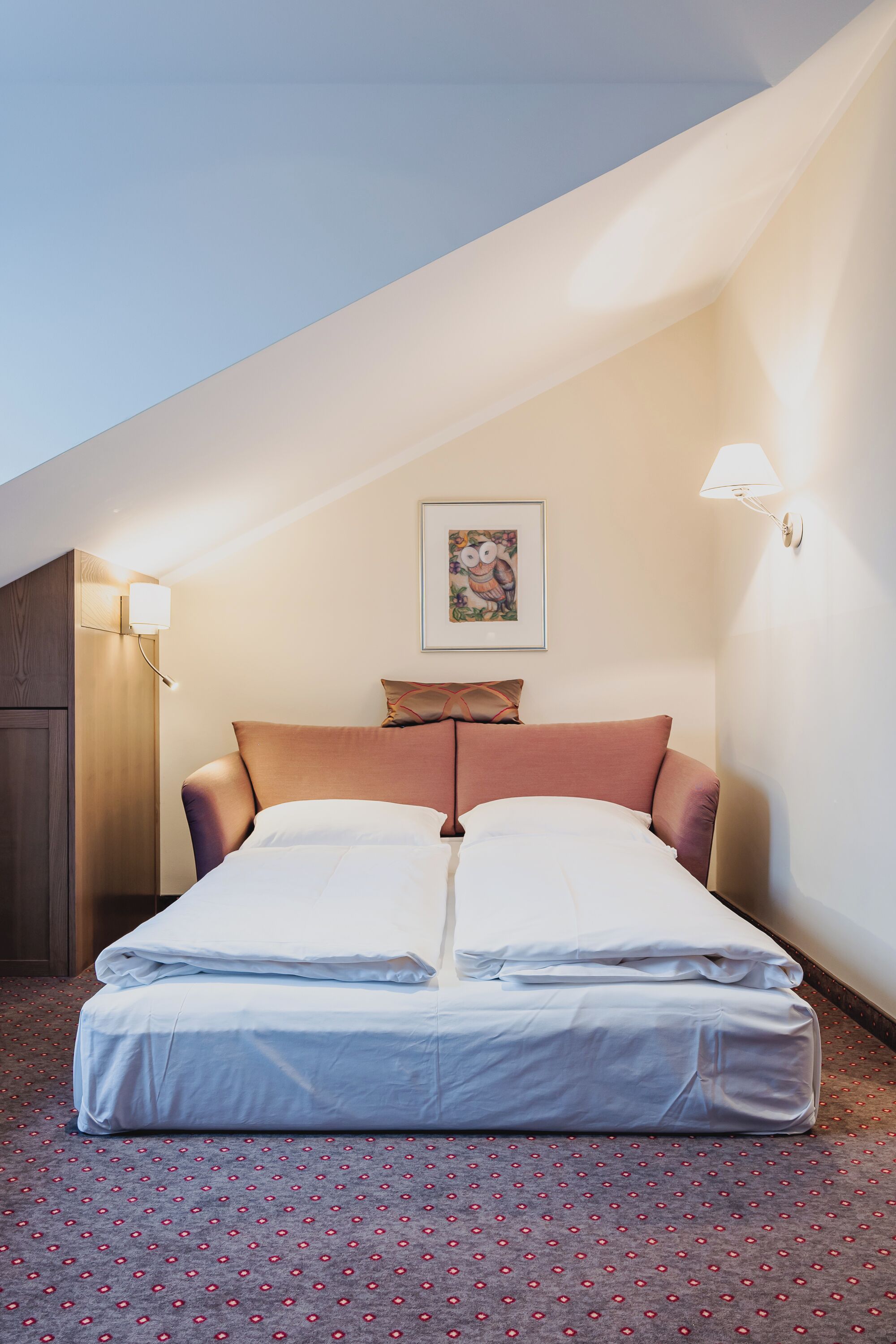 A neatly made bed in a room with a sloped ceiling, a framed picture on the wall, and a lamp on the side.
